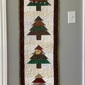 May include: A quilted wall hanging featuring a Christmas tree design in red, green and white. The tree is made from patchwork fabric and is set against a white background with a scrolling pattern. The hanging is framed with a red and green border.