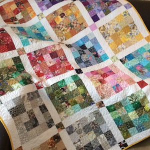 May include: A colourful patchwork quilt with a white background and a yellow border. The quilt is made up of many different squares of fabric, each with a unique pattern. The quilt is folded and draped over a brown sofa.