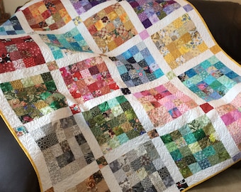 May include: A colorful patchwork quilt with a white background and a yellow border. The quilt is made up of many different squares of fabric, each with a unique pattern. The quilt is folded and draped over a brown couch.