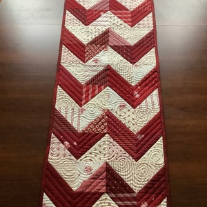 May include: A red and white chevron patterned table runner with a quilted design. The runner is made of fabric scraps in various patterns and colours.
