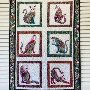 May include: A white quilt with a black paisley border and six colorful cat images in the center. Each cat is surrounded by a frame with a different color border. The cats are all in different poses and have a paisley pattern on their fur.