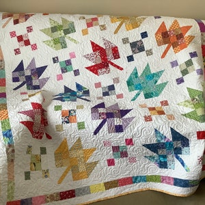 May include: A white quilt with a patchwork design featuring colourful maple leaves. The quilt is edged with a yellow border and a multi-coloured border.