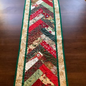 May include: A quilted table runner with a chevron pattern in red, green and white Christmas fabrics. The runner has a gold and green border.