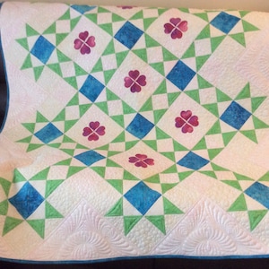 May include: A white quilt with a green and blue geometric pattern. The quilt is decorated with pink flower-shaped appliques.