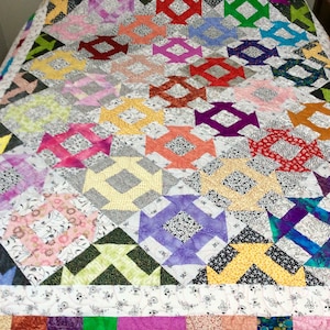 May include: A colourful patchwork quilt with a geometric pattern of squares and diamonds. The quilt is made of various fabrics in shades of pink, purple, red, yellow, green, and blue. The quilt is laid out on a bed with a wooden headboard.