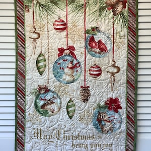 May include: A white quilted wall hanging with a green border and a Christmas theme. The design features red and white striped ornaments, pine branches, and a variety of other holiday decorations. The text "May Christmas bring you joy" is embroidered in gold on the bottom of the hanging.
