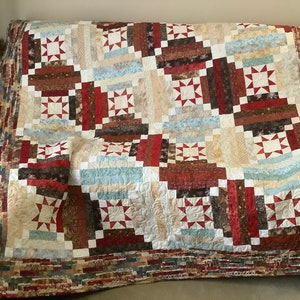 May include: A quilt with a log cabin pattern in shades of brown, red, beige, and blue. The quilt is made with a variety of fabrics and has a star design in the centre of each block.