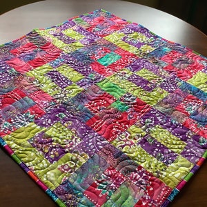 Peut inclure: Une courtepointe colorée en patchwork avec un schéma de couleurs vert, violet et rose. La courtepointe est faite de carrés de tissu cousus ensemble en un motif de grille. La courtepointe est matelassée avec un motif ondulé.