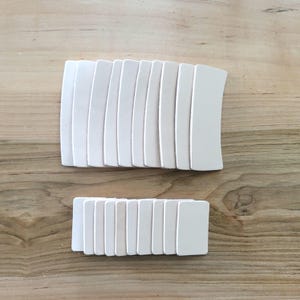 May include: Two sets of white, rectangular, curved-edge wooden pieces arranged on a light brown wooden surface. The larger set is curved, while the smaller set is rectangular.