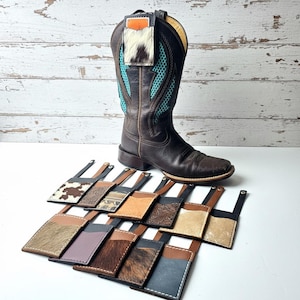 May include: A brown leather cowboy boot with turquoise detailing and a small leather tag. A selection of small leather wallets with diverse textures and patterns are arranged in front of the boot, displaying a range of designs.