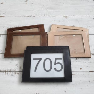 May include: A collection of leather house number plaques in various colors: brown, tan, and black. The black plaque displays the number "705" in large, bold white digits. Each plaque has a clear window for the number.