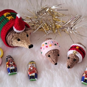 Puede incluir: Cuatro adornos de erizo tejidos con diferentes sombreros de temática navideña. Los adornos están rodeados de oropel dorado, cascanueces de chocolate y pequeñas monedas redondas de chocolate.