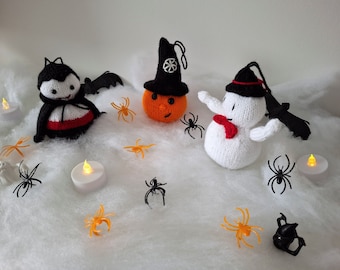 May include: A white knitted snowman with a black top hat and red bow tie, a black knitted vampire with red cape, and an orange knitted pumpkin with a black witch hat are surrounded by black and orange plastic spiders and white tea lights on a white fluffy surface.