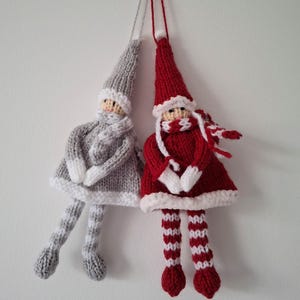 以下が含まれることがあります： 2つの編み物で作られたクリスマスのエルフオーナメント。1つは白のアクセントが付いたグレー、もう1つは白のアクセントが付いた赤です。どちらも長いとがった帽子、スカーフ、ストライプの脚が付いています。白い背景に吊り下げられています。