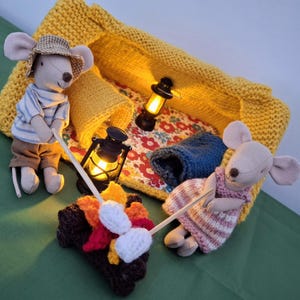 May include: A whimsical scene featuring two mouse dolls camping. One mouse wears a striped shirt and hat, the other a pink striped dress. A campfire with marshmallows, two lanterns, and a yellow knitted tent complete the scene.