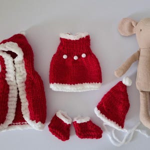 Op de afbeelding: Een beige stoffen muispop vergezeld van een set miniatuur rood-witte gebreide kleding, waaronder een cape met capuchon, een jurk met drie parelmoerachtige knopen, een paar kleine slofjes en een puntmuts met een witte pompon.