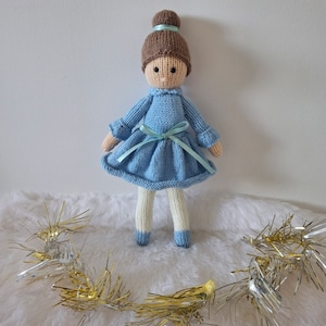May include: A knitted doll with brown hair in a bun, wearing a light blue dress with a light blue ribbon, white tights, and blue shoes. The doll is standing on a white fluffy surface with gold and silver tinsel.