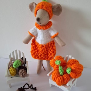 May include: A plush mouse toy wearing a crocheted orange pumpkin hat, a white and orange jumper, and a skirt. Beside it are two miniature white chairs, one holding crocheted acorns in green, yellow, and brown, and the other holding orange pumpkins.