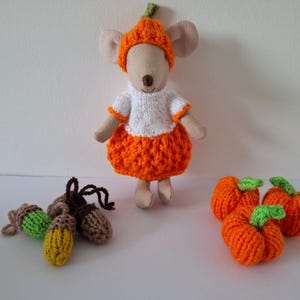 May include: A stuffed mouse toy wearing an orange pumpkin hat, a white short-sleeved top, and an orange skirt. Beside the mouse are three crocheted acorns in green, yellow, and brown, and three small crocheted orange pumpkins with green stems.