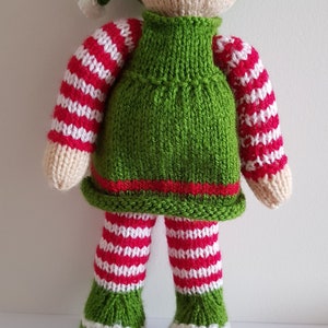 May include: A knitted elf doll with a green hat, red and white striped sleeves and pants, and green shoes. The elf has a smiling face with black eyes and a pink mouth.