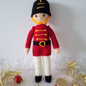 Puede incluir: Un soldado de juguete de punto que lleva un uniforme rojo y negro con un cinturón amarillo y un sombrero negro. El soldado lleva un pequeño tambor dorado.