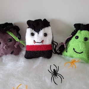 May include: Three knitted Halloween trick-or-treat bags with different character faces. The bags are purple, white and red, and green. The bags are sitting on a white fluffy surface with two black and orange spiders.