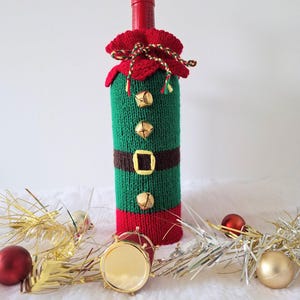 May include: A green and red knitted wine bottle cover designed to look like an elf. The cover has a red collar, a brown belt with a gold buckle, and three gold bells. The bottle's neck is adorned with a red cap and gold tinsel.