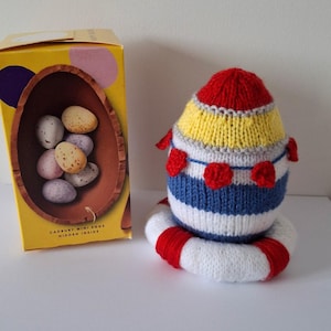 Op de afbeelding: Een gebreid paasei met rode, witte, blauwe en gele strepen. Het ei zit op een rode en witte gebreide ring. De doos op de achtergrond zegt "Cadbury Mini Eggs Hidden Inside".