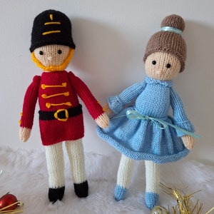 Puede incluir: Un muñeco de juguete de punto que representa a un cascanueces que lleva un uniforme rojo y negro con un cinturón amarillo y un sombrero negro.  Junto al cascanueces hay un muñeco de juguete de punto que representa a una bailarina que lleva un vestido azul claro con una cinta azul claro y un moño marrón.