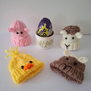 May include: Four knitted egg cozies in the shape of animal heads. A pink pig, a white sheep, a yellow chick, and a brown bear. A chocolate egg is in a white egg cup.