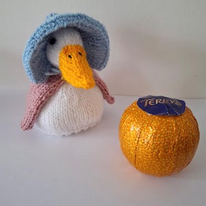 May include: A handmade knitted duck wearing a blue bonnet and pink shawl, with a yellow beak. Beside it is a Terry's chocolate orange wrapped in orange foil with a blue label.