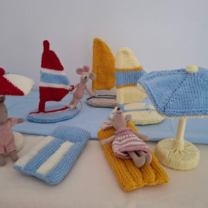 May include: A selection of knitted beach toys, featuring a red and white sailboat, a blue and white striped beach mat, a yellow and white beach umbrella, and several small mouse figures. The toys are in various colours, including red, yellow, blue, and white.