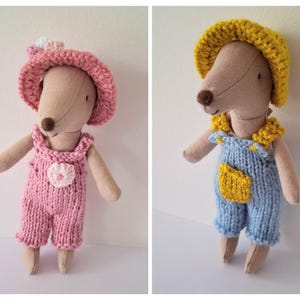 May include: Two handmade fabric dolls. One doll wears a pink knitted romper with a matching hat and a flower accent. The other doll is dressed in blue knitted dungarees and a yellow hat and collar. Both dolls have tan bodies and brown noses.