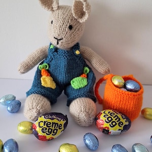May include: A knitted bunny wearing blue overalls with green and yellow accents. The bunny is holding a basket filled with chocolate eggs. There are several chocolate eggs scattered around the bunny.
