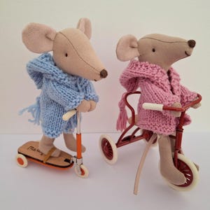 May include: Two beige fabric mouse dolls with stitched features. One mouse wears a light blue knit hooded sweater and stands on an orange and wood scooter. The other mouse wears a pink knit hooded sweater and sits on a red metal tricycle.