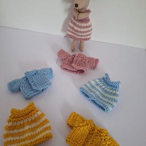 May include: A soft toy mouse in a pink and white striped dress, surrounded by miniature knitted garments. The garments include sweaters and tops in pink, blue, yellow, and white stripes. The mouse has a light brown body and large ears.