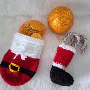 Könnte beinhalten: Ein gestrickter Beutel im Weihnachtsmann-Design mit weißem Umschlag, rotem Körper und schwarzem Gürtel mit gelber Schnalle, gefüllt mit goldfarbenen Münzen. Daneben ein kleiner gestrickter Stiefel-Anhänger mit zwei Zuckerstangen und einer goldenen Kugel.