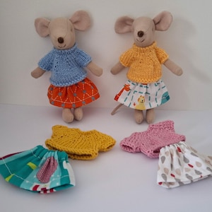May include: Two beige fabric mouse dolls standing upright. One wears a blue knit jumper and orange skirt, the other a yellow knit jumper and animal print skirt. Two additional outfits are laid out: a yellow knit top with a teal skirt and a pink knit top with a white skirt featuring a raindrop pattern.