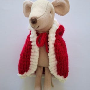 Puede incluir: Una muñeca de ratón de tela beige con nariz y orejas cosidas en marrón. El ratón lleva una capa de punto roja con un borde blanco difuso y un lazo de hilo rojo en el cuello. La muñeca tiene brazos y piernas largos y delgados.