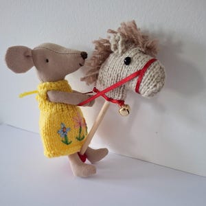 May include: A soft toy mouse in a yellow dress with floral embroidery, holding a wooden stick attached to a knitted horse's head. The horse has a red bridle and a small gold bell. The toys are set against a white background.