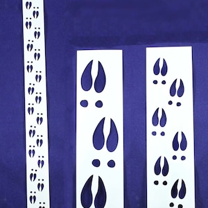 May include: Three white vinyl stencils with deer hoof prints. The stencils are different sizes and show multiple hoof prints. The largest stencil has two hoof prints. The medium stencil has three hoof prints. The smallest stencil has a row of 15 hoof prints.