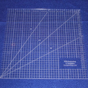 May include: A clear plastic ruler with a grid pattern and angled lines marked with 30, 45, and 60 degrees. The ruler is 12 inches by 12 inches and has a company logo in the bottom right corner that reads "TCR Engraving & Graphics, tcrengraving.com".
