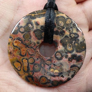 Puede incluir: Un colgante circular con un agujero central, con un patrón moteado de marrón, naranja, verde y negro. El colgante está suspendido de un cordón negro. La piedra tiene una superficie pulida.