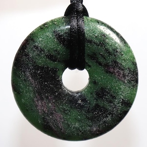 Può includere: Un ciondolo in pietra verde e nera di forma circolare con un foro centrale, sospeso a un cordino nero. La pietra ha un aspetto maculato. Un gioiello.