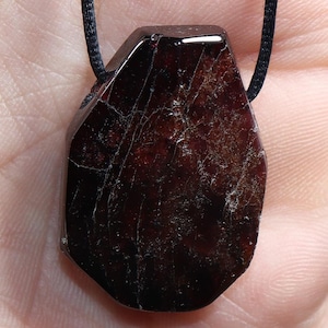 Puede incluir: Colgante de piedra preciosa facetada de color rojo oscuro con cordón negro. El colgante tiene una forma irregular con fracturas internas visibles. El color profundo de la gema le da un aspecto único y natural.