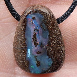 Könnte beinhalten: Ein tropfenförmiger Boulder-Opal-Anhänger mit einer dunkelbraunen, texturierten Matrix und einer zentralen Ader mit irisierenden blauen, grünen und roten Blitzen. Der polierte Stein hängt an einer einfachen schwarzen Kordelkette.