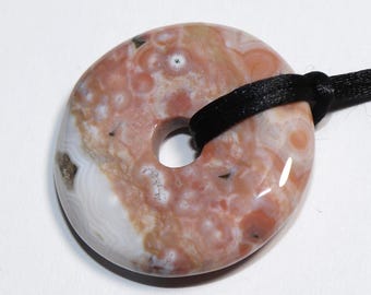May include: A polished, donut-shaped pendant necklace. The stone features a mix of pink, white, and brown hues with a central hole. A black cord is threaded through the pendant for wearing.