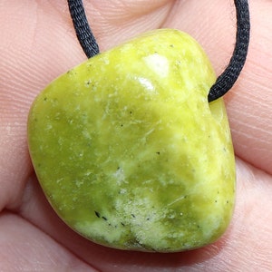 Peut inclure: Un pendentif en pierre arrondie vert clair, monté sur un cordon noir. La pierre présente un aspect moucheté avec des taches vertes plus foncées et noires. Le pendentif est tenu dans une main, mettant en valeur sa texture et sa couleur naturelles.