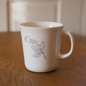 Könnte beinhalten: Weiße Keramiktasse mit einem grauen Rosenmuster. Die Tasse hat einen Henkel und steht auf einer Holzoberfläche.