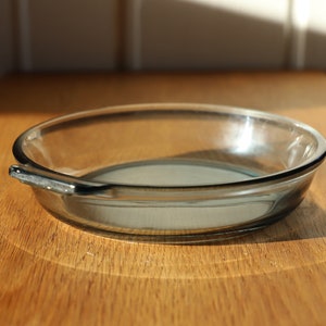 Può includere: Una pirofila di vetro trasparente con un'unica maniglia sul lato. La pirofila è appoggiata su una superficie di legno.